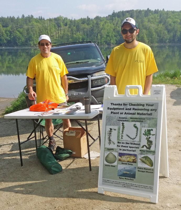 Two men wearing yellow shirts stand behind a table on the left and a sign on the right.  The sign reads "Thanks for Checking Your Equipment and Removing any Plant or Animal Materiall".  The sign has seven pictures each of different Aquatic Invasive Species on it.